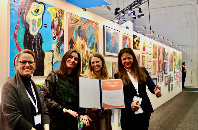 Vier junge Frauen sehr fröhlich und ausgelassen. Eine hält beide Daumen hoch. Sie halten die Urkunde / Auszeichnung in die Kamera. Sie stehen im Ausstellungsraum, hinter ihnen eine  lange Wand mit vielen Kunstwerken. Alle freue sich über die Urkunde.