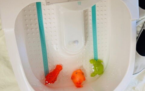 Ein Fußsprudelbad mit drei kleinen Quietschetieren. Davor hockt ein Kind mit weiteren Tieren neben sich. Auf dem Sprudelbad liegt eine Karte mit 6 Metacom Symbolen u.a. Wasser, Tiere, an, aus, rein und raus.