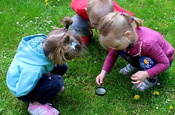Drei kleine Kinder, 2 Mädchen und ein Junge hocken auf einer Wiese und schauen durch eine Lupe, die das eine Mädchen über das Gras hält