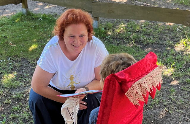 Kind sitzt auf kleinem roten Samt-Thron. Eine Frau hockt davor und lächelt fröhlich.