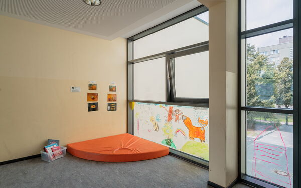 Ecke eines Raums. Eine orangene Matratze liegt in der Ecke, das untere Fenster der Ecke ist bamalt mit Fuchs, Biene, Regenwolke und Sonne.
