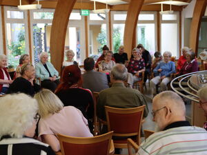 Großer Raum, größtenteils ältere Menschen sitzen im Halbkreisen auf Stühlen.