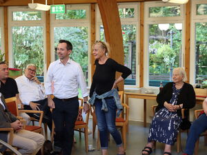 5 Menschen sitzen, ein Mann in weißem Hemd und eine Frau mit blonden Haaren und Brille auf den Kopf geschoben stehen und blicken nach links.