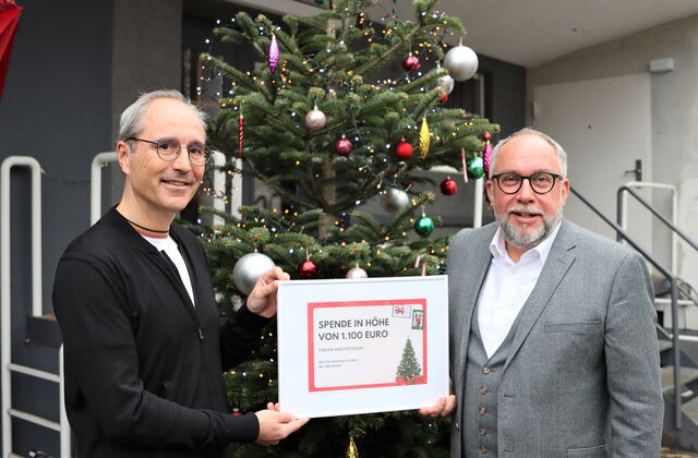 Zwei Männer um die 50 mit einer eingerahmten Spende. Text: Spende in Höhe von 1.100 Euro. Die Männer stehen draußen vor einen bunt geschmückten Weihnachtsbaum. Beide graue Haare, Brille, sind feierlich gekleidet grauer Anzug / weißes Hemd.
