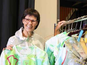 Ältere Frau mit kurzen dunklen Haaren, runder Brille mit dunklem Rahmen, hellgrauen Kapuzenpulli. Sie steht an einem Kleiderständer und hat die Hand auf die Stange gelegt. Am Kleiderständer hängen viele Kostüme und eines hält die Frau in die Hand.