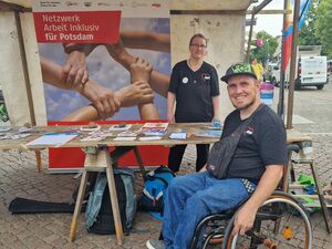 Ein Stand auf dem Fest mit einem Plakat von Netzwerk Arbeit inklusiv für Potsdam. Info-Material auf einem Tisch. Eine Frau im AWO T-Shirt steht hinter dem Tisch. Davor ein Rollstuhlfahrer im Aktivrollstuhl mit AWO T-Shirt und Base Cape.