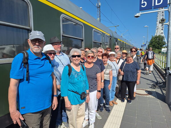 Gruppenbild: ältere Menschen auf einem Gleis. Der Zug steht im Bahnhof. Der Zug ist dunkelgrün. Es ist strahlend blauer Himmel.