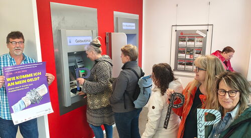 Menschen-Schlange vor Geldautomat. Einige Frauen halten ein großes P In die Kamera. Ein Mann steht links außen und hält ein lila Plakat "Wie komme ich an mein Geld?"