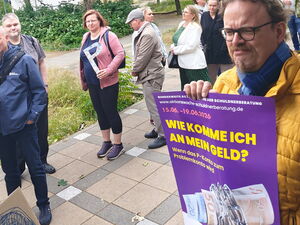 Draußen: Menschen-Schlange. Einige Personen halten ein großes P in der Hand.   Rechts im Bild Mann mit gelber Jacke hält lila Plakat vor sich.