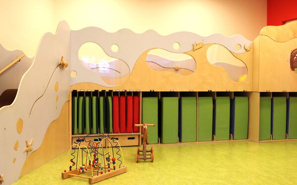 Teil eines hellen Raumes, der wie ein Spielbereich für Kinder gestaltet ist. Dominierend ist eine große, freistehende Holzeinrichtung, die an eine Wellenlandschaft erinnert.