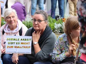 Drei Frauen sitzen. Um sie rum Protestierende. Die Frau in der Mitte schaut in die Kamera und hält ihr kleines weißes Protestschild in Richtung der Kamera. Text darauf: Soziale Arbeit gibt es nicht zum Nulltarif.