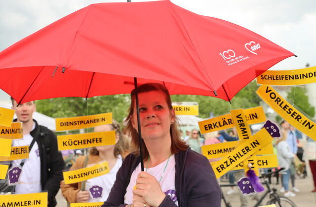 Frau mit großem roten AWO-Regenschirm. Im Hintergrund Menschen mit gelben Schildern. Text auf verschiedenen Schilder: Vorleser, Schleifenbinder, Erzähler Dienstplaner, Küchenfee
