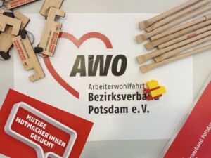 AWO-Schlüsselanhänger, AWO-Kugelschreiber. Das AWO Logo ist auf die Tischfläche geklebt und es liegt eine rote Karte auf dem Tisch mit Text: Mutige Mutmacher*innen gesucht.