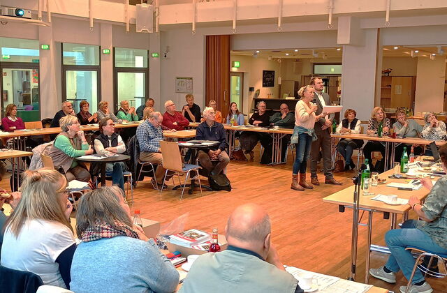 Sehr großer Raum, Tische zu einem großen Viereck gestellt an denen vielen Menschen sitzen. In deren Mitte stehen eine Frau und mit Mikro und ein Mann.