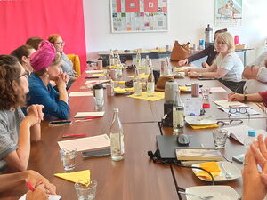 Großer Raum, große Tische. Viele Menschen sitzen sich gegenüber, einige machen sich Notizen.