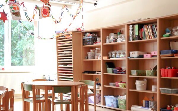 Großes Wandregal mit Bastelmaterialien. Ein Runder Tisch mit Stühlen. Von der Decke hängen gebastelte Strene und Girlanden und Bilder von Kindern.