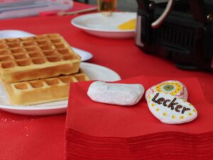 Zwei Waffeln auf einem Teller daneben liegt ein Stapel roter Servierten die mit zwei Steinen beschwert sind. Der eine Stein ist hübsch bemalt mit Blumen und dem Wort "Lecker".