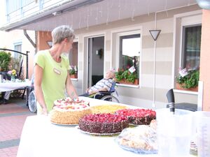 Draußen vor einem Haus. Ein Tisch mit 5 verschiedenen Torten und Kuchen. Eine Frau steht an dem Tisch und schaut zu einem alten Mann der im Rollstuhl am Haus sitzt.