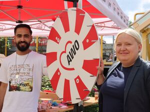 Ein Mann und eine Frau lächeln in die Kamera. Zwischen ihnen ein rot-weißes AWO Glücksrad. Der Mann mit schwarzen Haaren und Vollbart, schlank mit T-Shirt. Blonde Frau, etwas fülliger im dunklen Blazer.