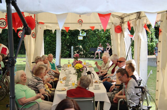 Draußen, Festzelt, mit rot-weißen Wimpel, roten und weißen Luftballons geschmückt. Lange Festtafel an der Senioren mit Kaffee und Kuchen sitzen, Das Wetter ist sonnig.