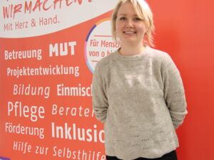 Portrait: Junge blonde Frau lächelt in die Kamera. Sie trägt einen beigen Pullover, eine schwarze Hose und hat die Hände hinter dem Rücken.