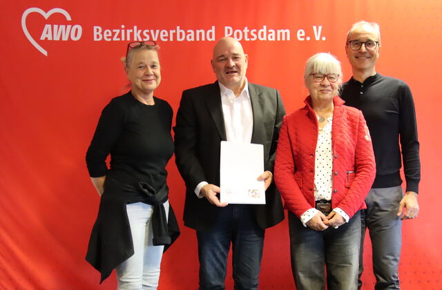 Portrait: 4 Personen vor roter AWO Wand. Frau, blonde lange Haare hochgesteckt, Brille auf den Kopf geschoben, Mann mit Glatze, weißem Hemd, schwarzem Sakko hält ein Papier vor sich, Frau mit weißen Haaren und Brille und großer schlanker Mann mit Brille.