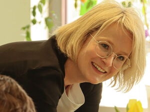 Kinder beim gemeinsamen Essen am Tisch. Am Tischende steht eine blonde Frau mit Brille die sich auf den Tisch lehnt und zur Kamera lächelt.