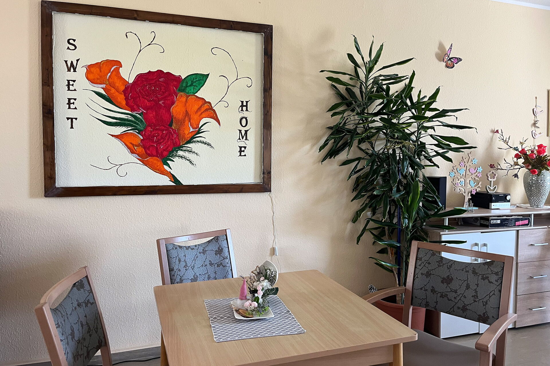 Gemeinschaftsraum: großes Bild mit Raosen und dem Text: SWEET HOME an der Wand. Davor ein Tisch um den drei Stühle stehen. Eine große Zimmerpflanze und ein Sideboard stehen an der Wand.