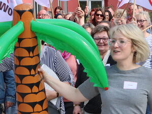Eine Zielflagge links im Bild. Eine blonde Frau mit einer großen Gummi-Palme tritt über die Ziellinie (rotes Band) Hinter hier Teilnehmende im weißen AWO Flaggen.
