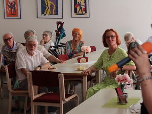 Raum mit Menschen an Tischen. Im Vordergrund rechts: Blonde Frau mit Mikro in der linken Hand.