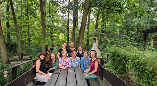 Gruppenfoto: 9 Personen sitzen im Halbrund an einem Holztisch, der in einem bewaldeten Gelände steht. Alle sehen in die Kamera.
