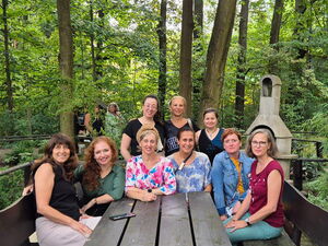 Gruppenfoto: 9 Personen sitzen im Halbrund an einem Holztisch, der in einem bewaldeten Gelände steht. Alle sehen in die Kamera.