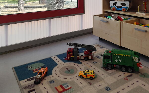 Teppich mit einem Straßenmuster, darauf sind Spielzeugautos und -figuren platziert. Im Hintergrund ist ein Holzregal mit Spielzeug und Pflanzen sowie ein Fenster, durch das Tageslicht hereinfällt.