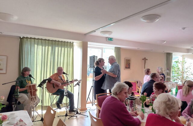 Heller Raum mit großen Fenstern. Kaffeetafeln und zwei Menschen die am Fenster sitzen und Musik machen. Rechts von dem Musikern ein tanzendes Paar.