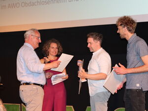 Preisübergabe. Älterer Mann übergibt Urkunden an Sieder. Zwei Männer nehmen Pris entgegen. Moderatorin lächelt.