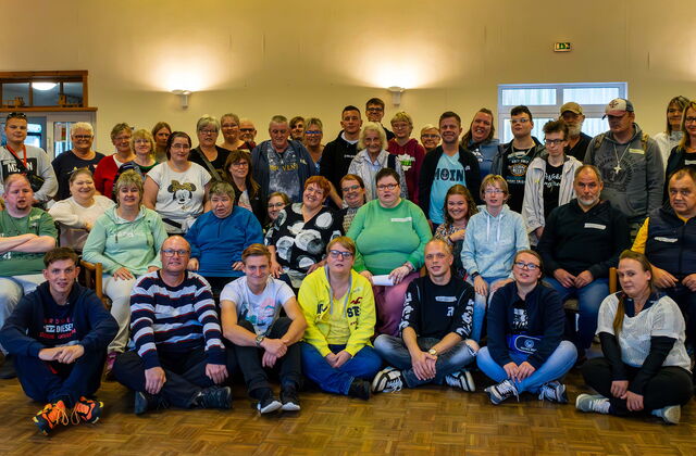 Großes Gruppenfoto in einer großen Halle. Vorne sitzen Menschen auf dem Fußboden, dahinter ist eine Reihe Stühle, ein Rollstuhlfahrer steht auch außen, dahinter stehen noch viele weitere Menschen.