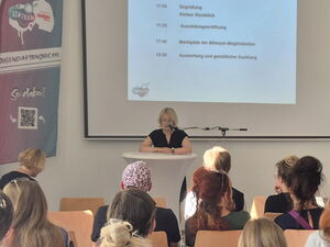 Publikum auf Stühlen von hinten. Am Pult eine blonde Frau die in ein Mikrofon spricht. An einer Projektionswand der Ablaufplan des Tages.