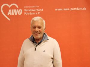 Portrait: Älterer Mann mit weißen Haaren und Drei-Tage-Bart. Er trägt einen weißen Pullover und lächelt in die Kamera. Hinter ihm eine orangene AWO Wand mit Logo und Website-Adresse awo-potsdam.de