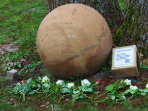 Draußen an einem Baumstamm ist eine Gedenkkugel. Davor liegen Blumen. Die Kugel besteht aus rot gebranntem Ton mit einer Inschrift:. Die Kugel hat eine kupferne Färbung.