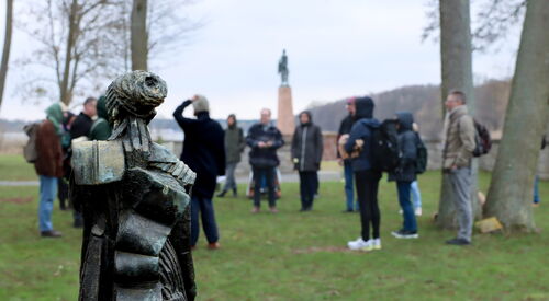 Im Vordergrund ein Denkmal. Dahinter draußen auf einer Wiese zwischen Bäumen stehen einige Menschen in einem Kreis.