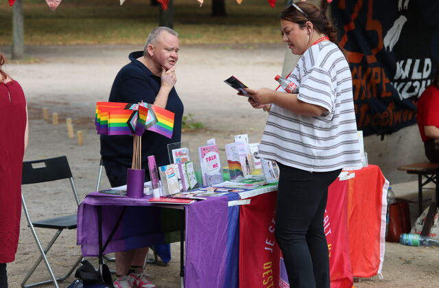 Älterer Mann an einem Rogenbogen-buntem Stand mit Büchern und Info-Material informiert eine interessierte Frau.
