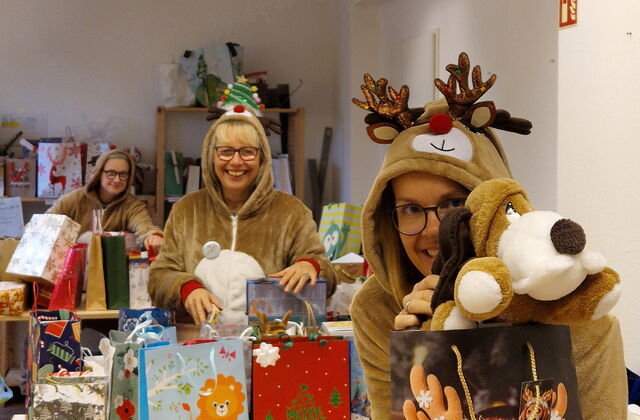 Gefüllte Geschenktüten. Ganz votn mit einem Weihnachtshund drauf. Frau im Rentierkostüm hält einen Plüschhund darüber und lächelt. Dahinter noch eine Frau im Rentierkostüm mit mehreren Geschenktüten. Alle sind sehr fröhlich.