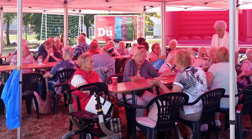 Tische und Stühle unter roten AWO Pavillons. Ältere Menschen sitzen beisammen und unterhalten sich.