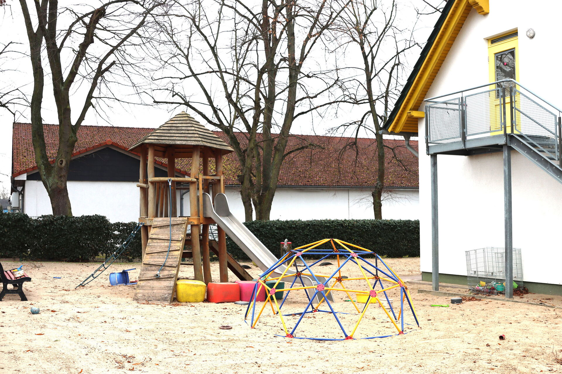 Spielplatz mit Krettergerüst mit Rutsche und einer Bank. Umgeben von einer Hecke. Daneben weißes Haus mit einer Außentreppe die zu einer gelben Tür im Obergeschoss des Hauses führt.