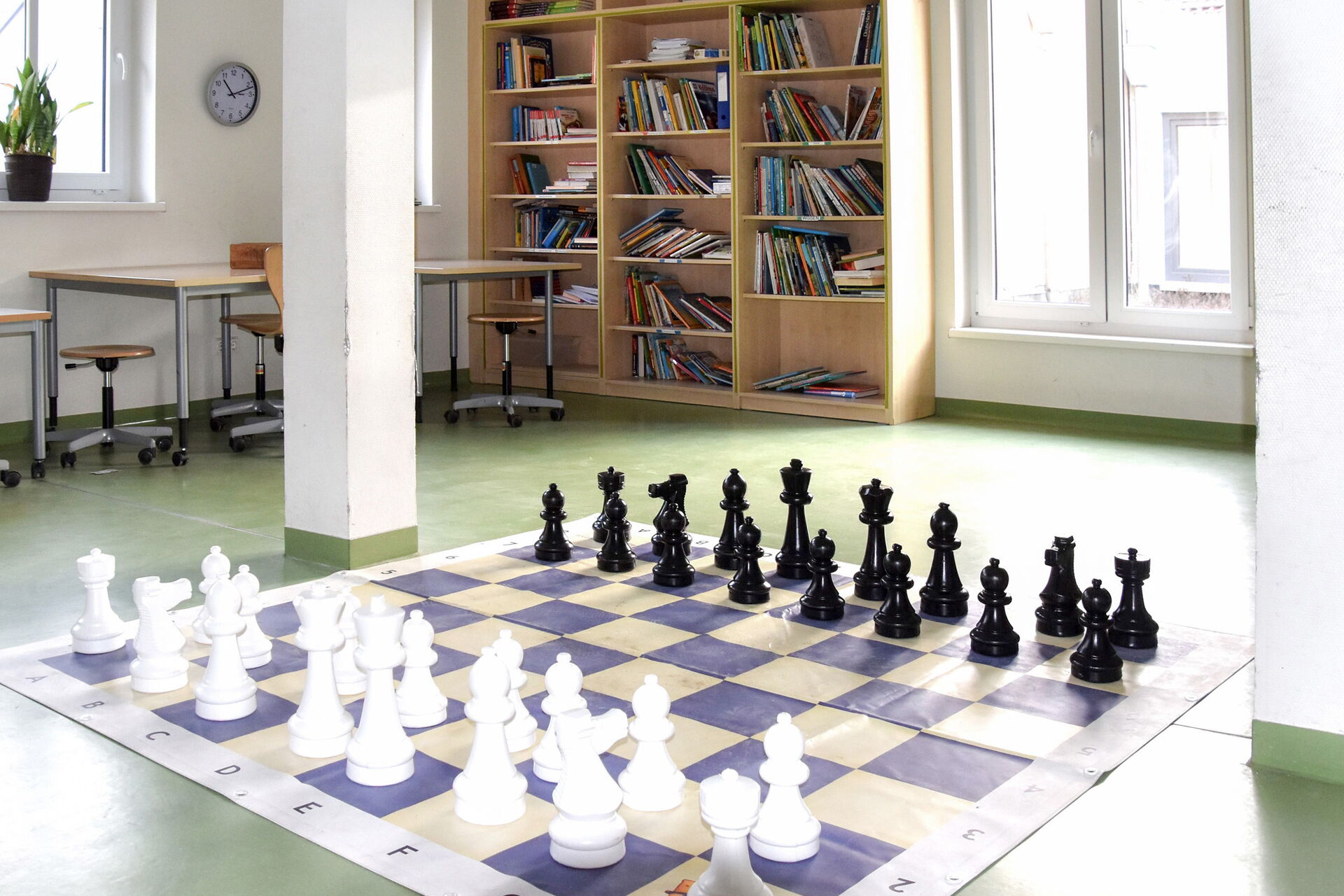 Ein Spielraum. Ein großes Schachspiel ist auf dem Fußboden aufgebaut. In der Zimmerecke ein Bücherregal mit Büchern.