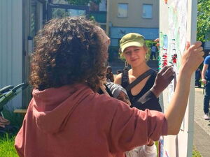 2 junge Menschen halten ein Pinntafel, währen eine Person einen Pin anpinnt. Draußen, Sonne scheint.