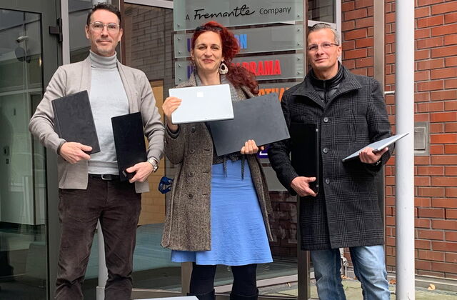 2 Männer in der Mitte eine Frau, alle drei halten Laptops und Notebooks in den den Händen. Sie stehen vor dem Gebäude der UFA.