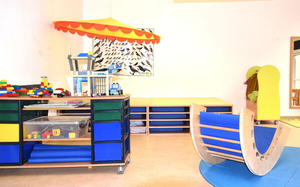 Spielzimmer. Kleines Regal mit Lego. Kleine halbrunde Wippe aus Holz mit blauer Polsterung innen. An der hinteren Wand ein Schaubild mit verschiedenen Vögeln.