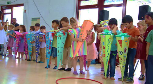 Ca. 12 Einschulungskinder mit ihren großen Zuckertüten in einem großen Raum. Die Zuckertüten sind farbig und bund beklebt. Die Kinder freuen sich.