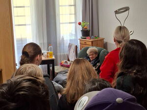 Bewohnerzimmer. Seniorin sitzt im grünen Sessel Schüler und eine Mitarbeiterin stehen mit dem Rücken zur Kamera.